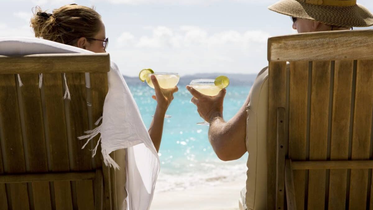 people sit in two wooden beach chairs facing the Caribbean ocean holding drinks and making a toast